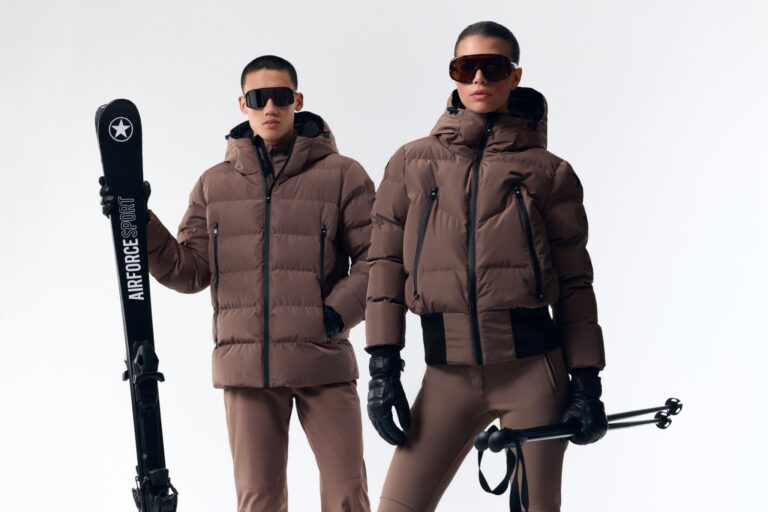 Heren en dames model in matchende bruine Airforce Sport ski-outfits met ski's en stokken, verkrijgbaar bij Sportshop Pier.